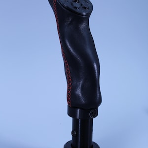 Puede incluir: Un pomo de palanca de cambios de cuero negro con costuras rojas. El pomo tiene un agarre texturizado y una base de metal negro. La parte superior del pomo tiene un diseño estampado. Accesorio automotriz.