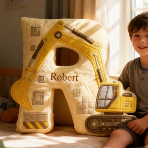 Op de afbeelding: Een decoratief kussen in de vorm van de letter 'R' met een gele graafmachine. Het kussen is crèmekleurig en heeft de naam 'Robert' erop geborduurd. De graafmachine heeft gele en grijze details.