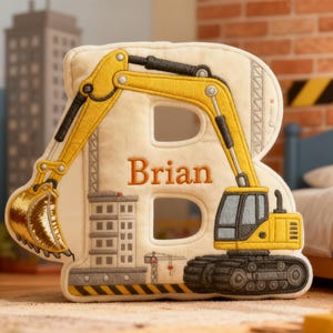 Op de afbeelding: Een crèmekleurig kussen in de vorm van de letter 'B' met een gele graafmachine. De naam 'Brian' is oranje geborduurd. De graafmachine heeft een zwarte arm en bak, met een bouwplaats.