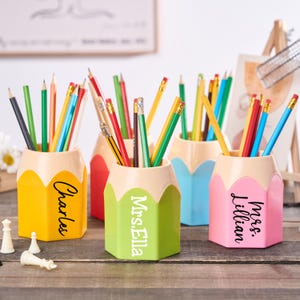 May include: A selection of brightly coloured pencil-shaped desk tidies, each holding pencils. The organisers are yellow, red, green, blue, and pink, with names printed on the sides. Chess pieces are in the foreground.