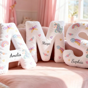 Puede incluir: Cojines de letras de felpa blanca que deletrean "AMS" con unicornios, arcoíris y mariposas bordados. La letra "A" tiene escrito "Amelia", "M" tiene "Maya" y "S" tiene "Sophia". Los cojines están sobre un sofá rosa.
