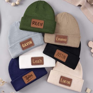 Può includere: Una collezione di berretti personalizzati in vari colori, tra cui verde, blu, nero e beige. Ogni berretto presenta una toppa rettangolare in pelle con un nome, come "Riley" o "Gavin". I cappelli sono disposti su una superficie grigio chiaro.