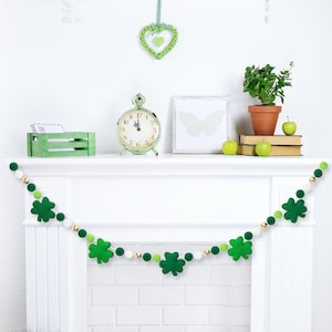 以下が含まれることがあります： 白い暖炉のマントルの上に、白いポンポンが付いた緑のクローバーのガーランドが飾られています。ガーランドは、緑のクローバーと白いポンポンで飾られています。暖炉のマントルは、緑の時計、白い額縁、茶色の鉢に入った緑の植物で飾られています。