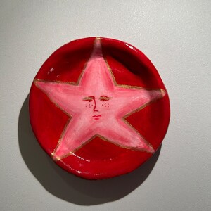 May include: A red ceramic dish with a pink star painted on the inside. The star has a face with closed eyes and a small mouth.