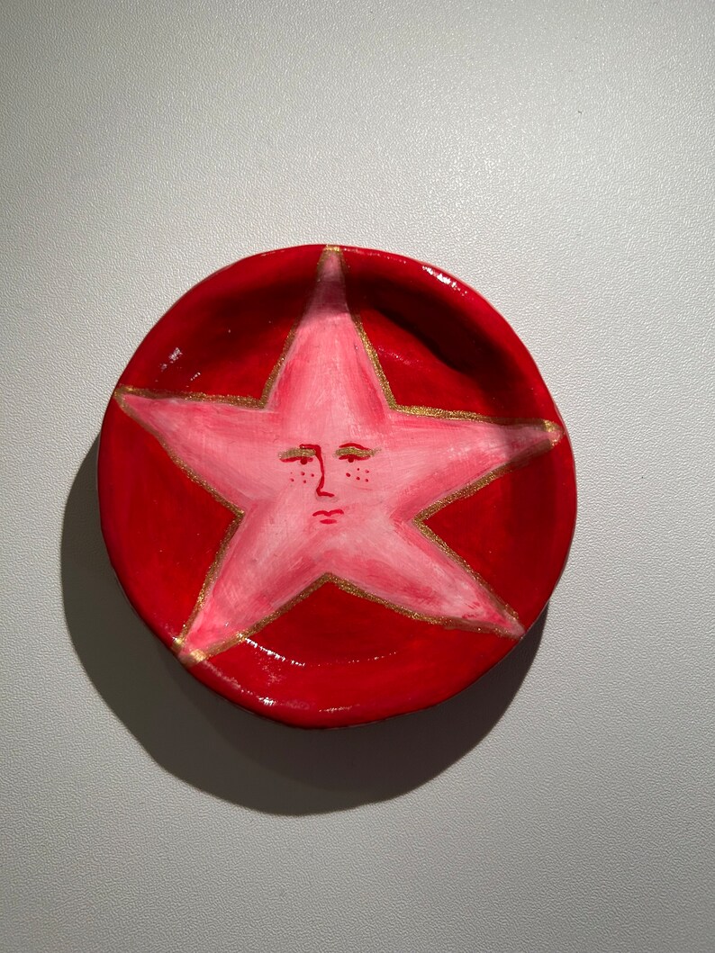 May include: A red ceramic dish with a pink star painted on the inside. The star has a face with closed eyes and a small mouth.
