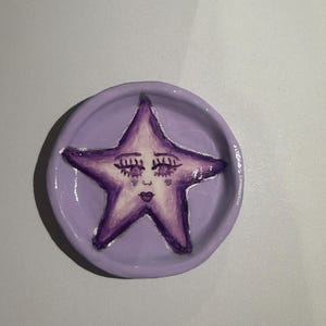 May include: A purple ceramic dish with a hand-painted purple star with a face. The star has closed eyes, a nose, and a mouth.