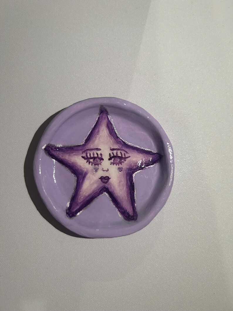 May include: A purple ceramic dish with a hand-painted purple star with a face. The star has closed eyes, a nose, and a mouth.
