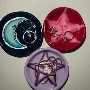 May include: Three ceramic ring dishes with painted designs. One dish is blue with a crescent moon, one is red with a pink star, and one is purple with a purple star. Each dish has silver rings inside.