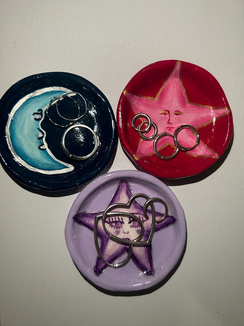 May include: Three ceramic ring dishes with painted designs. One dish is blue with a crescent moon, one is red with a pink star, and one is purple with a purple star. Each dish has silver rings inside.