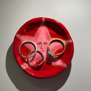 May include: A red ceramic ring dish with a pink star design and gold accents. Three rings are resting on the dish, two silver and one gold.