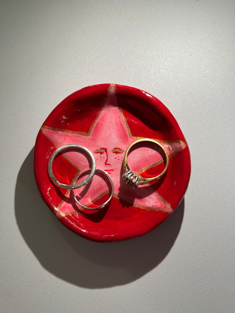 May include: A red ceramic ring dish with a pink star design and gold accents. Three rings are resting on the dish, two silver and one gold.