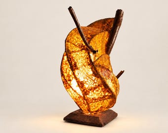 May include: A table lamp featuring a glowing, amber-colored, textured shade with a swirling, organic shape. The shade is supported by dark, branch-like wooden elements and rests on a square, dark brown wooden base.