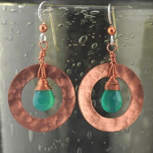May include: A pair of copper earrings with green gemstone drops. The earrings are hammered and have a circular shape with a smaller circle in the center. The gemstones are suspended from the smaller circle.