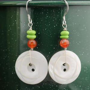 May include: A pair of handmade earrings featuring white, button-shaped pendants with a spiral design. The earrings have a silver wire and are accented with green and orange beads. The background is a dark green.