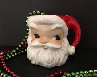 May include: A vintage ceramic Santa Claus mug with a red hat and white beard. The mug has a winking eye and a cheerful expression.