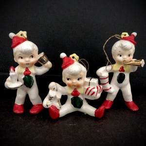 Könnte beinhalten: Drei weiße Porzellan-Weihnachtsornamente, die Elfen darstellen, die rote und weiße Hüte tragen. Die Elfen halten eine Kerze, einen Zuckerstock und ein Musikinstrument.