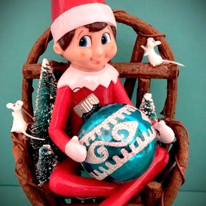 May include: A red and white elf figurine sitting in a wooden chair, holding a blue and silver Christmas ornament. The elf is wearing a red hat and a red and white suit. There are two white mice figurines on the chair.
