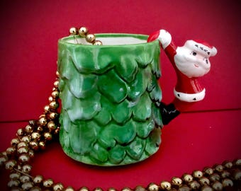 May include: A green ceramic Christmas tree mug with a Santa Claus handle. The mug is decorated with a textured tree design and a gold bead garland. The Santa handle is red, white, and black.