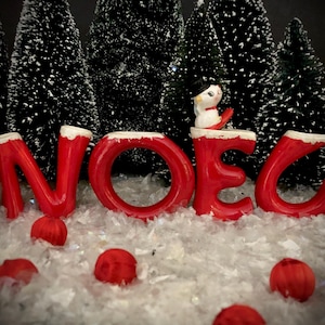 Könnte beinhalten: Rote Keramikbuchstaben, die "NOEL" buchstabieren, mit einer weißen Schneemannfigur, die auf dem "E" sitzt. Die Buchstaben stehen auf einer weißen, schneebedeckten Oberfläche mit roten Ornamenten, die verstreut sind.