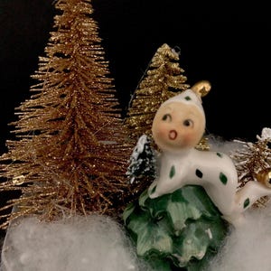 May include: A white ceramic figurine of a whimsical character with a green and white spotted outfit, sitting on a green pine cone. The figurine is surrounded by two gold glitter pine trees and white cotton-like snow.