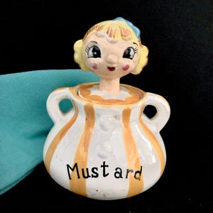 Pode incluir: Um pote de mostarda vintage de cerâmica com um design listrado amarelo e branco. O pote tem duas alças e a palavra "Mostarda" escrita na frente. A tampa apresenta uma menina caprichosa com cabelo loiro e um laço azul.