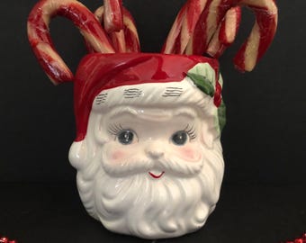 May include: A ceramic Santa Claus head-shaped container, painted in white, red, and flesh tones, filled with red and white striped candy canes. The container has a red hat and a white beard. A strand of red beads is in the foreground.