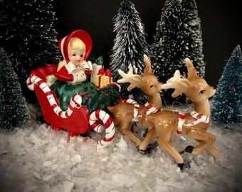 May include: A vintage Christmas figurine featuring a girl in a red sleigh pulled by three reindeer. The sleigh has candy cane accents, and the girl is wearing a red hood and green dress. The scene is set in a snowy landscape with miniature Christmas trees.