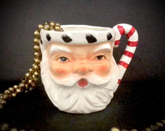 May include: A white ceramic Santa Claus mug with a red and white candy cane handle. The mug has a black and white striped hat and a white beard.