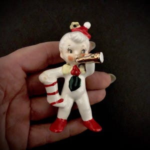 Könnte beinhalten: Ein vintage Porzellan-Weihnachtsornament, das einen Jungen zeigt, der eine rot-weiße Mütze und einen weißen Anzug mit rot-weißen Streifen trägt. Er hält eine goldene Trompete an seinen Mund und trägt eine grüne Fliege.