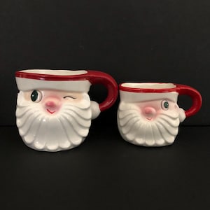 Puede incluir: Dos tazas de cerámica de Papá Noel. Las tazas son blancas con detalles rojos en el asa y el borde. La cara de Papá Noel está pintada en la parte delantera, con un guiño en una taza. Ideales para bebidas navideñas.