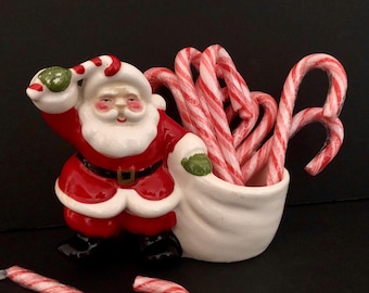 May include: A ceramic Santa Claus figurine in a red suit with a white beard and hat, holding a white bag filled with red and white striped candy canes. A candy cane lies in front of the figurine. Festive holiday decor.
