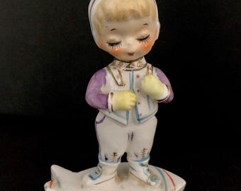 May include: A porcelain figurine of a child wearing a purple and white ski suit with a white hat and blonde hair. The child is standing on a white, star-shaped base.