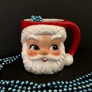 Könnte beinhalten: Eine Keramik-Weihnachtsmann-Tasse mit rotem Henkel und Hut, weißem Bart und Besatz sowie einem lächelnden Gesicht. Die Tasse ist mit blauen Perlen im Inneren und um die Tasse herum verziert.