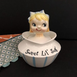 Puede incluir: Un dispensador de azúcar de cerámica blanca con rayas azules y las palabras "Sweet Lil' Sug" en la parte delantera. El dispensador tiene una cabeza de niña con cabello rubio y ojos azules en la parte superior.