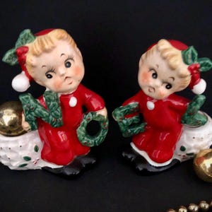 May include: Two vintage Christmas figurines. Each figurine is a small child in a red outfit, holding a green wreath and a gold ball. The figurines are on a white base with green and red accents. The figurines are approximately 4 inches tall.