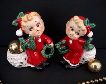 May include: Two vintage Christmas figurines. Each figurine is a small child in a red outfit, holding a green wreath and a gold ball. The figurines are on a white base with green and red accents. The figurines are approximately 4 inches tall.