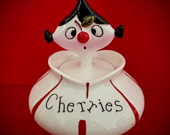 May include: A white ceramic jar with a whimsical design, featuring a black-haired figure with a cherry nose and a cherry on top. The word "Cherries" is written on the front in black script. Red accents and a red background complete the design.