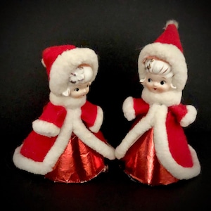 May include: Two vintage Christmas tree ornaments, each depicting a girl wearing a red and white dress with a white fur trim. The girls have white porcelain faces with painted features and black hair.