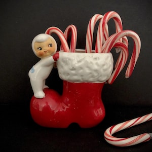 Könnte beinhalten: Eine Keramik-Weihnachtsstrumpf-förmige Süßigkeitenschale. Der rote Strumpf hat eine weiße, flauschige Verzierung und ist mit rot-weiß gestreiften Zuckerstangen gefüllt. Eine kleine weiße Figur mit lächelndem Gesicht lehnt sich an den Strumpf.