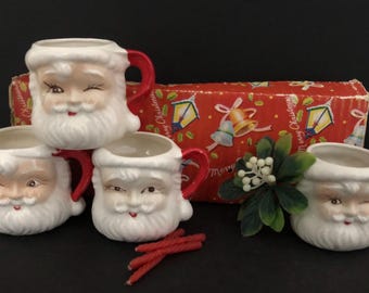 May include: Set of four ceramic Santa Claus mugs with red handles. Each mug features a winking Santa face with a white beard and rosy cheeks. A red and white patterned box is in the background, along with red candle sticks and a sprig of white berries.