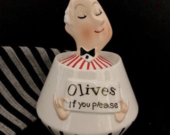 May include: A white ceramic olive dish shaped like a person with a red and white striped shirt, black bow tie, and a sign that reads "Olives If you please".