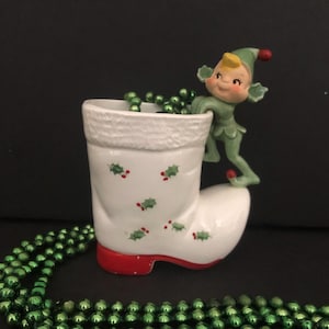 Könnte beinhalten: Ein weißer Weihnachtsstrumpf aus Keramik mit roter Basis und grünen Stechpalmenmustern. Eine grüne Elfenfigur sitzt auf dem Rand des Strumpfes.