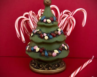 May include: A green ceramic Christmas tree with colorful ornaments and a gold base. Candy canes with red and white stripes surround the tree, with one lying in front. The background is a solid red.