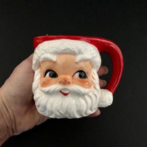 Könnte beinhalten: Eine Keramik-Weihnachtsmann-Tasse mit rotem Henkel und Hut. Die Tasse zeigt ein lächelndes Weihnachtsmann-Gesicht mit rosigen Wangen, einem weißen Bart und schwarzen Augen. Ein festlicher Artikel.