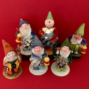 May include: A collection of vintage gnome figurines. Each gnome has a unique design, featuring cone-shaped bodies, fabric hats, and holding lanterns or small trees. The gnomes are set against a red background.