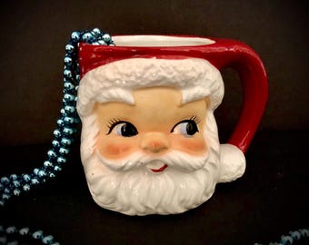May include: A vintage ceramic Santa Claus mug with a red hat and white beard. The mug has a handle and is decorated with a smiling face.