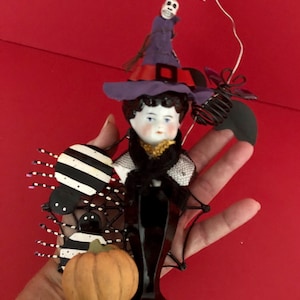 May include: A Halloween-themed figurine featuring a doll head with a witch hat, black dress, and spider accents. A small pumpkin is also included in the design. The figurine is held against a red background.