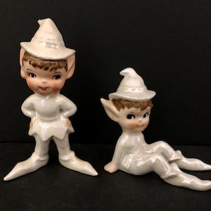 May include: Two ceramic elf figurines. One stands with hands on hips, the other sits with legs extended. Both wear white outfits and hats with brown hair peeking out. The figurines have rosy cheeks and large eyes.