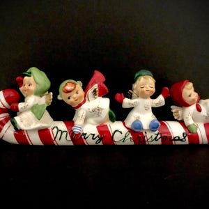 Könnte beinhalten: Eine antike Weihnachtsdekoration mit vier Porzellanengeln, die auf einem Zuckerrohr reiten, auf dem die Worte "Merry Christmas" stehen. Die Engel tragen weiße Kleider mit goldenen Akzenten und haben rote, grüne und blaue Akzente.