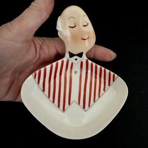 May include: A vintage ceramic ashtray shaped like a waiter's torso. The ashtray features a smiling face with closed eyes, a black bow tie, and a red and white striped shirt. The base is a cream color.